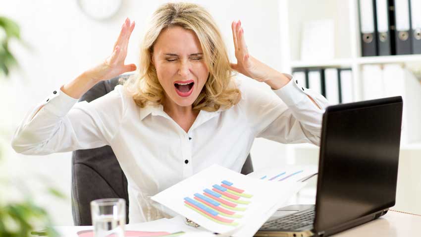 A stressed out woman seated at her office desk, throwing her hands up in frustration after experiencing holiday blues
